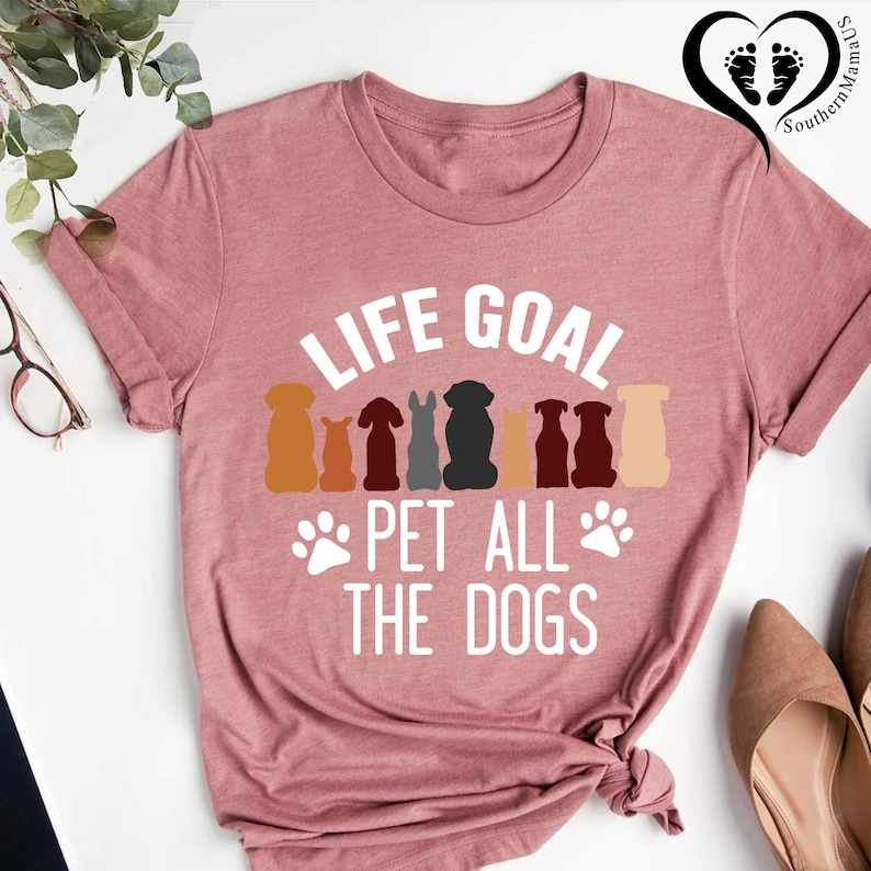 May include: Dusty rose t-shirt with the text "LIFE GOAL PET ALL THE DOGS" in white. Above the text are silhouettes of various dog breeds in shades of brown, gray, and black. Paw prints are below the text.
