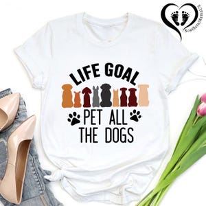 May include: White t-shirt with the text "LIFE GOAL PET ALL THE DOGS" in black. Above the text are silhouettes of various dog breeds in shades of brown, gray, and black. Two black paw prints flank the text.