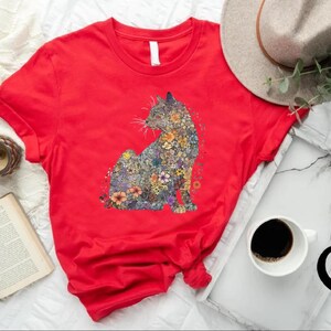 May include: A red t-shirt featuring a floral cat design. The cat is composed of various colorful flowers. The shirt is displayed with a hat, coffee, a book, and a clock. The Southern Mama's logo is visible.