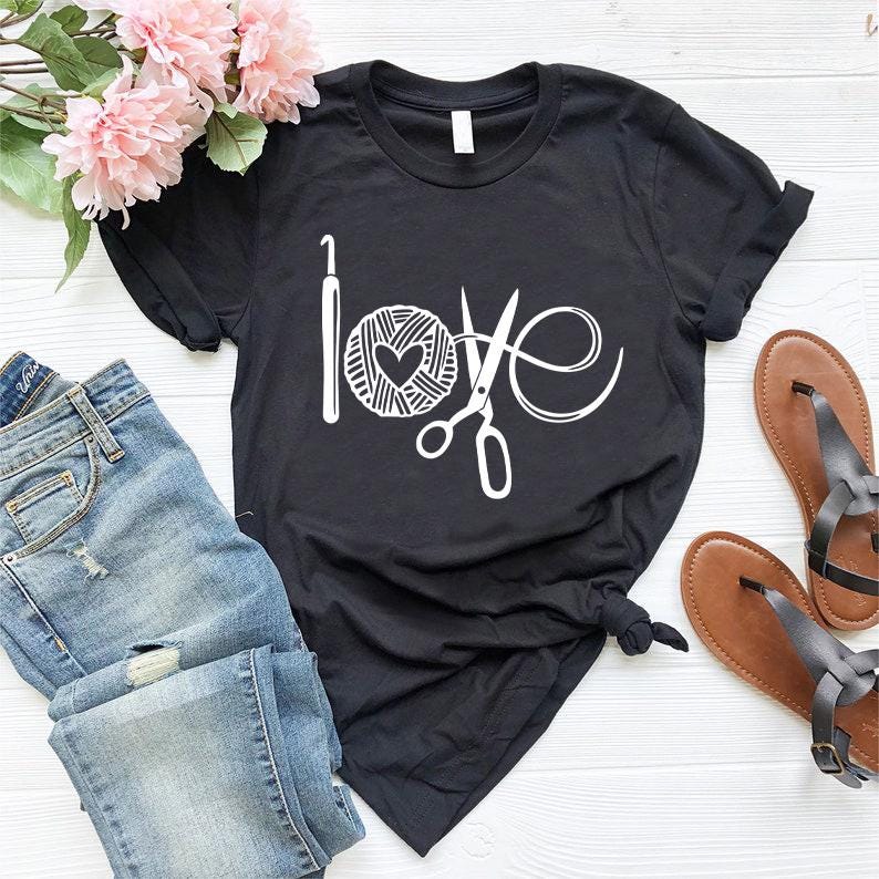 May include: Black t-shirt with a white graphic design that spells "love" using a crochet hook, yarn ball with a heart, and scissors. The shirt is paired with blue jeans, sandals, and pink flowers.