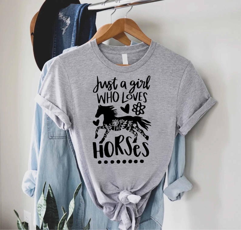 May include: A heather gray t-shirt with the text "Just a girl who loves horses" in black script. A black horse silhouette with floral detailing is centered below the text. The shirt is hanging on a wooden hanger.