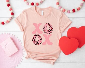 May include: A light pink t-shirt with the letters "XO" in a pink and leopard print design. Two red heart-shaped decorations are on the right side of the shirt. A pink gift box and a pink placemat are on the left side.