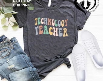 May include: A dark gray t-shirt with the words "TECHNOLOGY TEACHER" in colorful, retro-style lettering. The shirt is paired with blue jeans and white sneakers, suggesting a casual, everyday outfit. The image also includes a plant and a logo.