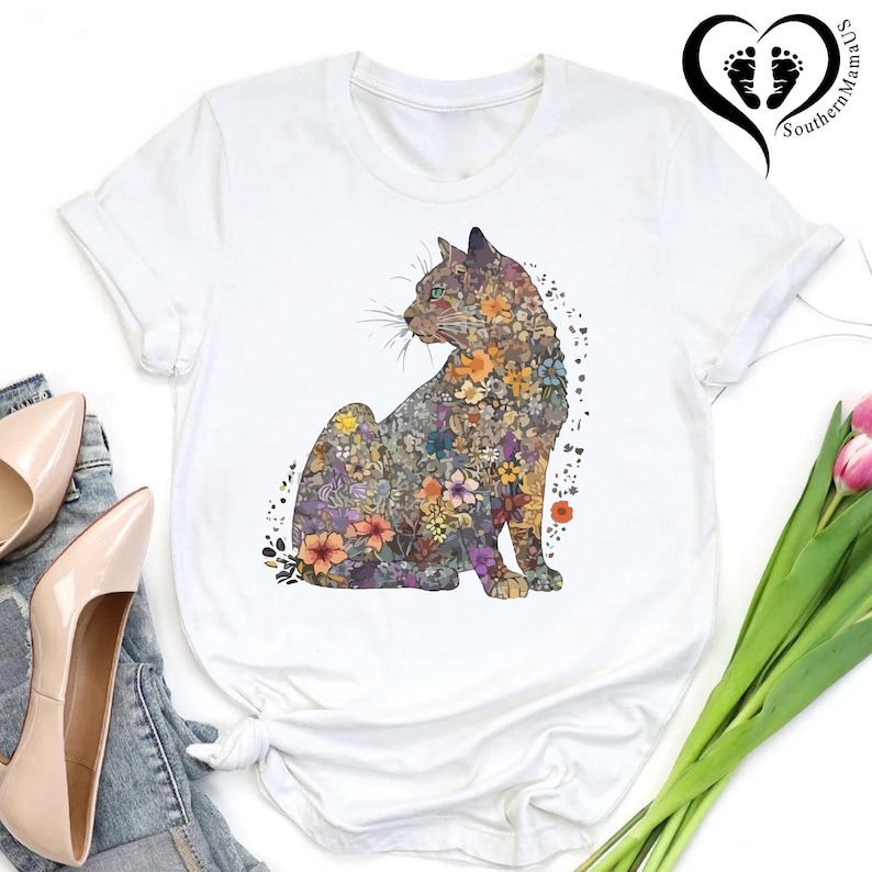 May include: White t-shirt featuring a seated cat design filled with colorful flowers. The cat illustration includes shades of purple, orange, yellow, and green. The shirt has short sleeves and a round neckline. The Southern Mama US logo is visible in the upper right corner.
