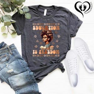 Puede incluir: Camiseta gris oscuro con las palabras "EDUCATION IS FREEDOM" en letras marrones y naranjas. La camiseta presenta una ilustraci&oacute;n de una mujer con gafas y libros. La camiseta se combina con vaqueros azules y zapatillas blancas.