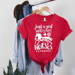 May include: A red t-shirt with the text "Just a girl WHO LOVES HORSES" in white script. A white horse graphic with floral details is below the text. The shirt is hanging on a clothing rack.