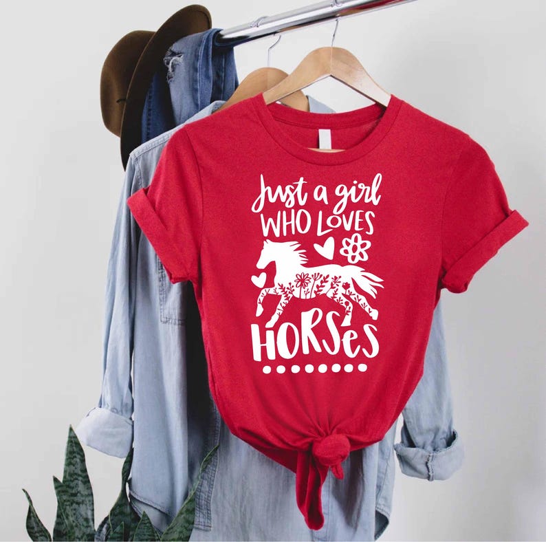 May include: A red t-shirt with the text "Just a girl WHO LOVES HORSES" in white script. A white horse graphic with floral details is below the text. The shirt is hanging on a clothing rack.