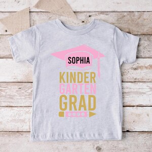May include: A light gray t-shirt with the name "SOPHIA" above the words "KINDERGARTEN GRAD". The text is in pink and gold colors, with a pink graduation cap graphic. A pencil graphic is at the bottom.