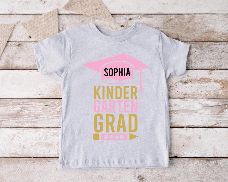 May include: A light gray t-shirt with the name "SOPHIA" above the words "KINDERGARTEN GRAD". The text is in pink and gold colors, with a pink graduation cap graphic. A pencil graphic is at the bottom.