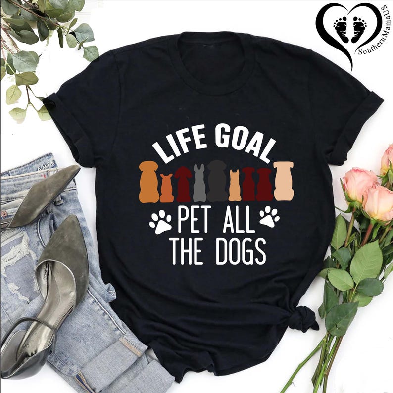 May include: Black t-shirt with the text "LIFE GOAL PET ALL THE DOGS" in white. The shirt features silhouettes of various dog breeds in brown, red, and gray. A pair of jeans and shoes are visible in the background.