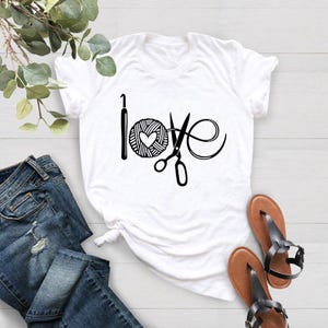 May include: White t-shirt with a black graphic that says "love" using a crochet hook, yarn ball with a heart, and scissors. The shirt is paired with blue jeans and brown sandals.