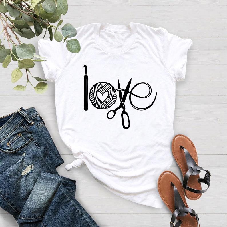 May include: White t-shirt with a black graphic that says "love" using a crochet hook, yarn ball with a heart, and scissors. The shirt is paired with blue jeans and brown sandals.