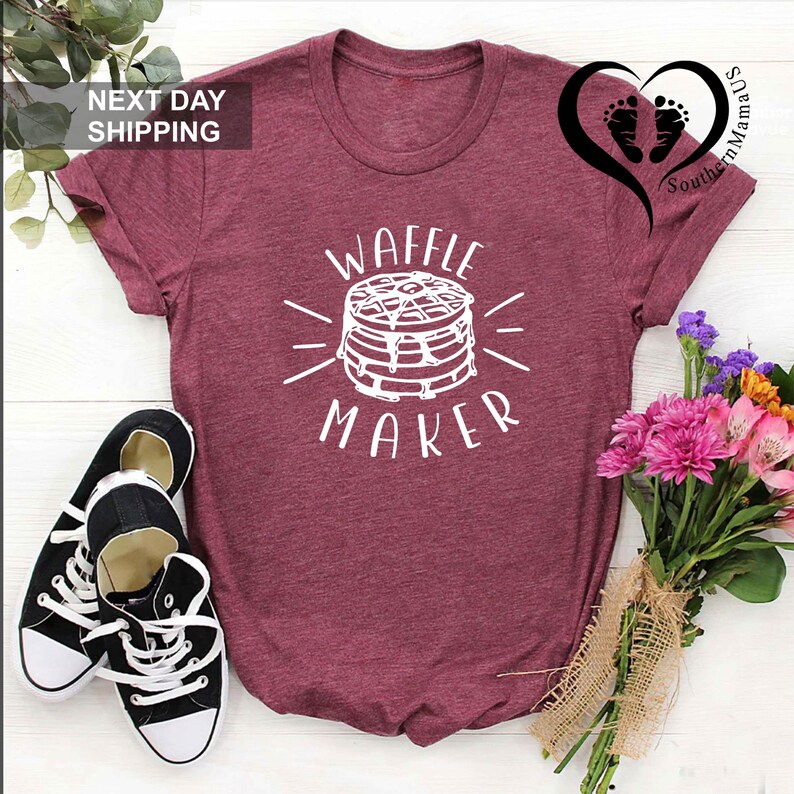 Puede incluir: Camiseta burdeos con las palabras "WAFFLE MAKER" y un gr&aacute;fico de gofres en blanco. Zapatillas deportivas blancas y negras est&aacute;n al lado de la camiseta. Un ramo de flores est&aacute; en el lado derecho de la imagen.