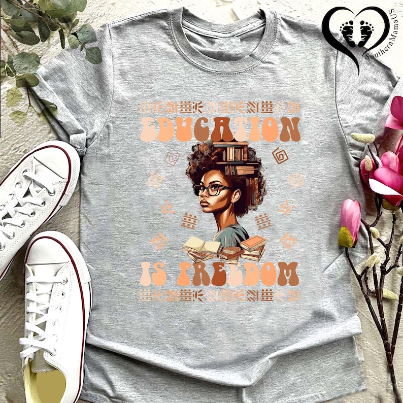 Puede incluir: Camiseta gris con las palabras "EDUCATION IS FREEDOM" en una fuente retro. La camiseta presenta una ilustraci&oacute;n de una mujer con gafas y libros en la cabeza. Se ven zapatillas blancas.