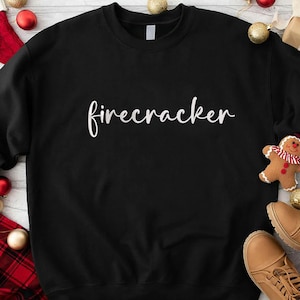 May include: Black sweatshirt with the word "firecracker" in white script. The sweatshirt is surrounded by Christmas decorations, including ornaments, a gingerbread man, and boots.