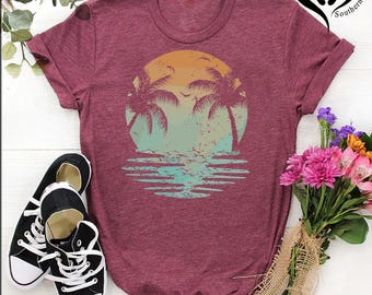 May include: Burgundy t-shirt featuring a sunset design with palm trees and water reflections. The graphic is in shades of orange, teal, and blue. Black and white sneakers are next to the shirt. A bouquet of flowers is on the right.