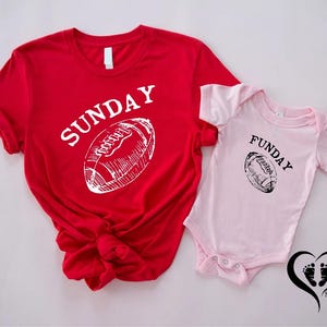 Puede incluir: Una camiseta roja con la palabra "SUNDAY" y un gr&aacute;fico de f&uacute;tbol americano blanco. Junto a ella, un body rosa para beb&eacute; con la palabra "FUNDAY" y un gr&aacute;fico de f&uacute;tbol americano negro. La imagen incluye un coraz&oacute;n con huellas.