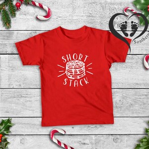 Puede incluir: Una camiseta roja con las palabras "Short Stack" en blanco y un gr&aacute;fico de panqueques. La camiseta se muestra sobre una superficie de madera blanca, decorada con elementos navide&ntilde;os como bastones de caramelo y acebo.