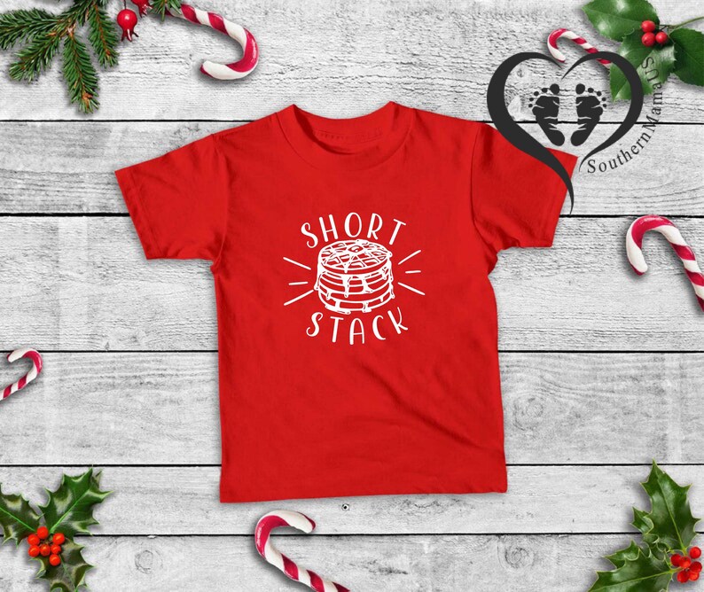 Puede incluir: Una camiseta roja con las palabras "Short Stack" en blanco y un gr&aacute;fico de panqueques. La camiseta se muestra sobre una superficie de madera blanca, decorada con elementos navide&ntilde;os como bastones de caramelo y acebo.