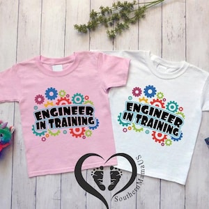 May include: Two short-sleeved t-shirts, one pink and one white, with the text "ENGINEER IN TRAINING" in a black font. The text is surrounded by colorful gear graphics. A plush toy and a small box are also visible.