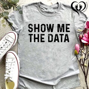 May include: A heather gray t-shirt with the phrase "SHOW ME THE DATA" printed in bold black letters. White canvas sneakers with red trim are visible on the left. The shirt is a casual, everyday wear item.
