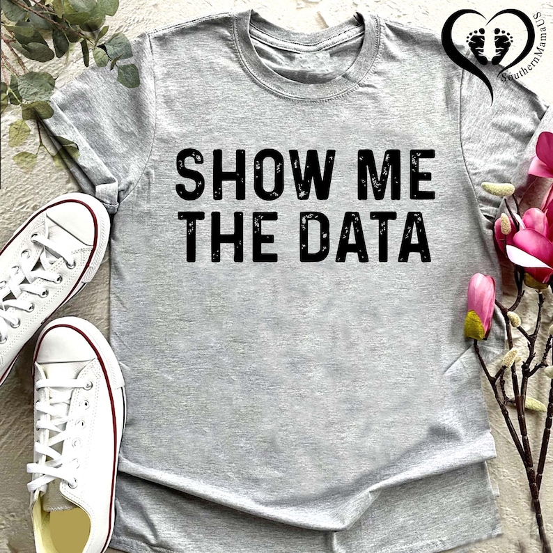 May include: A heather gray t-shirt with the phrase "SHOW ME THE DATA" printed in bold black letters. White canvas sneakers with red trim are visible on the left. The shirt is a casual, everyday wear item.