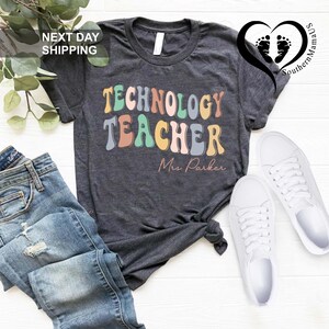 May include: A dark heather gray short-sleeve t-shirt featuring the text "TECHNOLOGY TEACHER" in a multi-colored, wavy, retro-style font, with the name "Mrs. Parker" printed below in a cursive script, paired with blue denim jeans and white sneakers.
