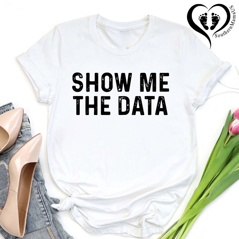 May include: White t-shirt with the text "SHOW ME THE DATA" in bold black letters. The shirt is styled with a knot at the bottom and is paired with light-colored heels and denim. A logo with footprints is in the upper right corner.