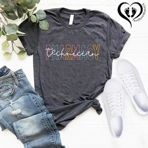 K&ouml;nnte beinhalten: Dunkelgraues T-Shirt mit der Aufschrift "Pharmacy Technician" in einer farbenfrohen, umrandeten Schriftart. Das Shirt wird mit blauen Jeans und wei&szlig;en Turnschuhen kombiniert. Ein Zweig mit Gr&uuml;n ist in der oberen linken Ecke zu sehen.