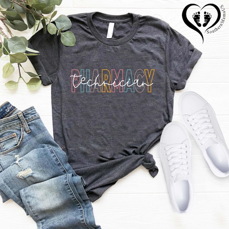 K&ouml;nnte beinhalten: Dunkelgraues T-Shirt mit der Aufschrift "Pharmacy Technician" in einer farbenfrohen, umrandeten Schriftart. Das Shirt wird mit blauen Jeans und wei&szlig;en Turnschuhen kombiniert. Ein Zweig mit Gr&uuml;n ist in der oberen linken Ecke zu sehen.