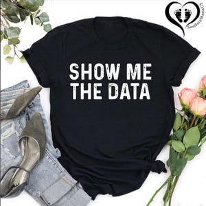 May include: Black t-shirt with the white text "SHOW ME THE DATA". The shirt is styled with a pair of blue jeans, gray heels, and a bouquet of pink roses. The Southern MamaUS logo is in the upper right corner.
