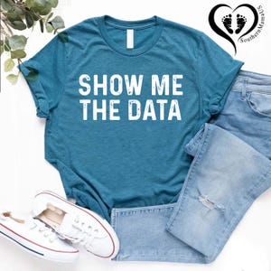 May include: Teal t-shirt with the text "SHOW ME THE DATA" in white block letters. The shirt is paired with light blue denim jeans and white sneakers with red and blue accents. A black heart-shaped logo with footprints is in the upper right corner.