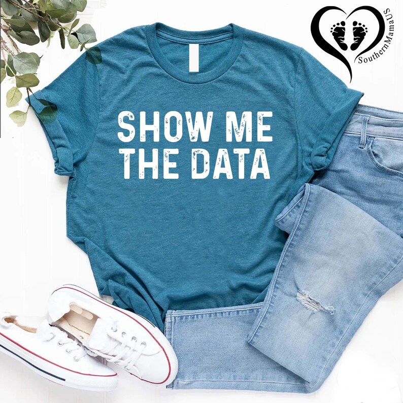 May include: Teal t-shirt with the text "SHOW ME THE DATA" in white block letters. The shirt is paired with light blue denim jeans and white sneakers with red and blue accents. A black heart-shaped logo with footprints is in the upper right corner.
