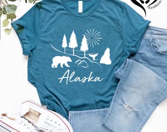 May include: Teal t-shirt with a white graphic of a bear, trees, sun, and whale tail, with the word "Alaska" in a script font. The shirt is paired with light blue jeans and white sneakers.
