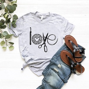 May include: A light gray t-shirt with the word "love" spelled out using a crochet hook, yarn ball with a heart, and scissors. The shirt is paired with blue jeans and brown sandals.