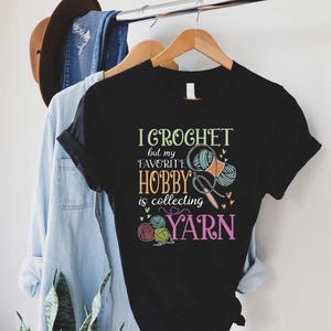 May include: Black t-shirt with the text "I Crochet but my favorite hobby is collecting yarn." The graphic includes yarn balls, scissors, and a crochet hook. A light blue denim shirt and jeans hang in the background.