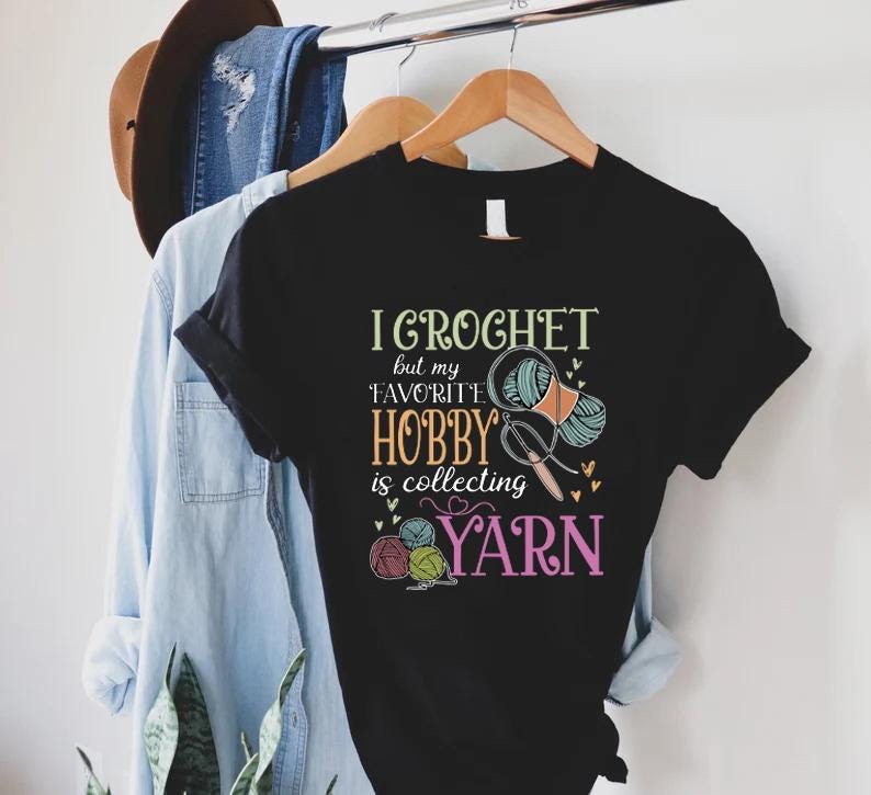 May include: Black t-shirt with the text "I Crochet but my favorite hobby is collecting yarn." The graphic includes yarn balls, scissors, and a crochet hook. A light blue denim shirt and jeans hang in the background.