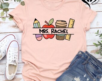 May include: A light pink t-shirt featuring a colorful graphic design for a teacher. The design includes a pencil, apple, coffee cup, and ruler, with the name "MRS. RACHEL" in the center. The shirt is paired with denim shorts and white sneakers.