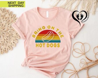 May include: A light pink t-shirt with the text "BRING ON THE HOT DOGS" in yellow. The text is above a graphic of a hot dog and a rainbow-colored background. The shirt has short sleeves and a crew neck.