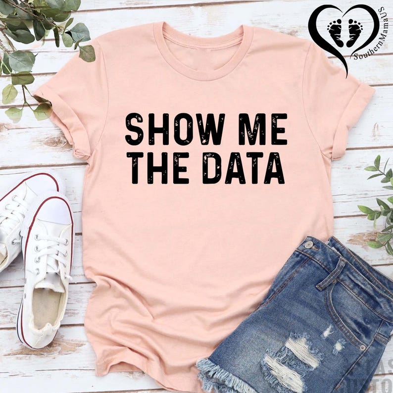 May include: A light pink t-shirt with the text "SHOW ME THE DATA" in bold black letters. The shirt is paired with denim shorts and white sneakers. The image is on a light wood background.