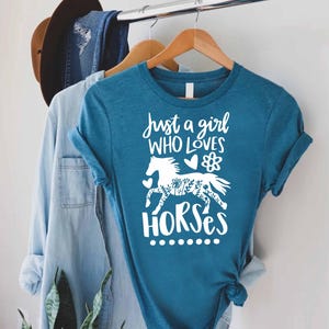 May include: Teal t-shirt with the text "Just a girl who loves horses" in white script. The design includes a white horse silhouette with floral details. The shirt is hanging on a wooden hanger.