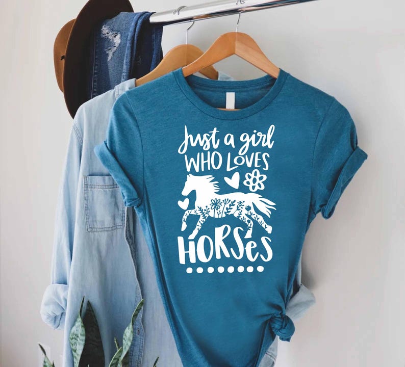 May include: Teal t-shirt with the text "Just a girl who loves horses" in white script. The design includes a white horse silhouette with floral details. The shirt is hanging on a wooden hanger.