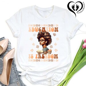 Puede incluir: Camiseta blanca con un gr&aacute;fico de una mujer con gafas y libros, con las palabras "EDUCATION IS FREEDOM" en marr&oacute;n y naranja. El dise&ntilde;o incluye patrones decorativos. La camiseta se combina con tacones beige y vaqueros.