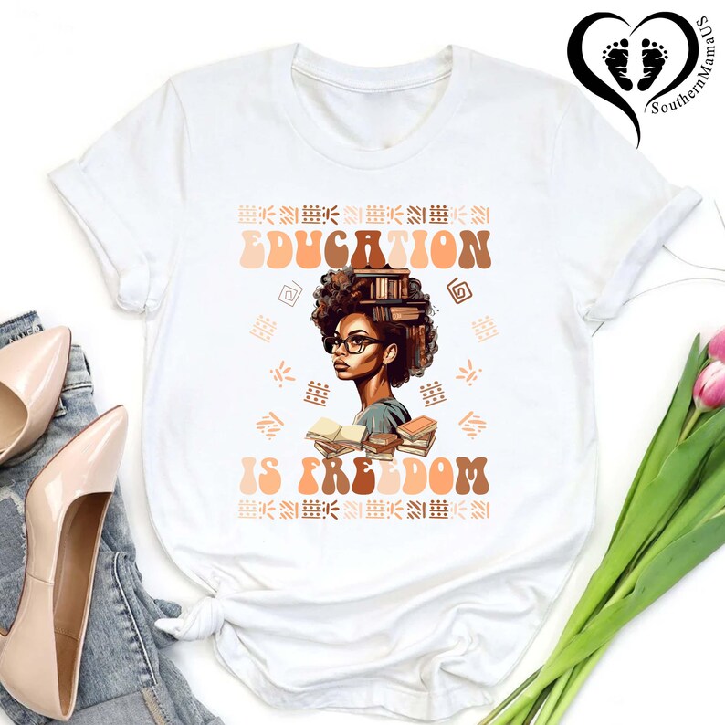 Puede incluir: Camiseta blanca con un gr&aacute;fico de una mujer con gafas y libros, con las palabras "EDUCATION IS FREEDOM" en marr&oacute;n y naranja. El dise&ntilde;o incluye patrones decorativos. La camiseta se combina con tacones beige y vaqueros.