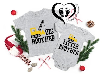 May include: Two gray shirts, one a short-sleeved tee and the other a onesie, feature a yellow construction vehicle graphic and the words "BIG BROTHER" and "LITTLE BROTHER" in black. Festive holiday decorations surround the shirts.