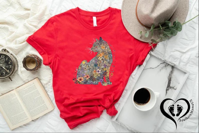 May include: A red t-shirt featuring a floral cat design. The cat is composed of various colorful flowers. The shirt is displayed with a hat, coffee, a book, and a clock. The Southern Mama's logo is visible.