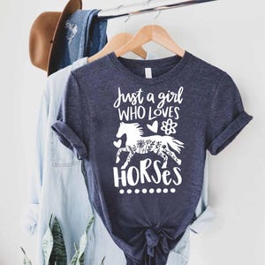 May include: Navy blue t-shirt with the text "Just a girl who loves horses" and a white graphic of a horse decorated with floral patterns. The shirt is hanging on a wooden hanger with other clothing.