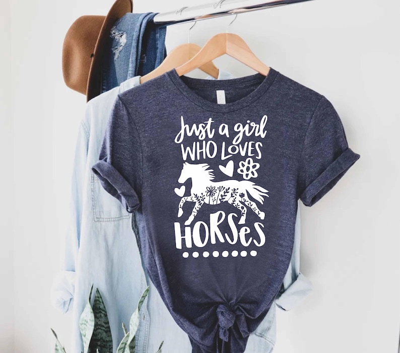 May include: Navy blue t-shirt with the text "Just a girl who loves horses" and a white graphic of a horse decorated with floral patterns. The shirt is hanging on a wooden hanger with other clothing.
