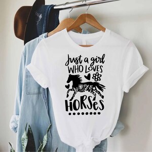 May include: White t-shirt with the text "Just a girl WHO LOVES HORSES" and a black horse graphic. The horse graphic has a floral design on its body. The shirt is hanging on a wooden hanger.