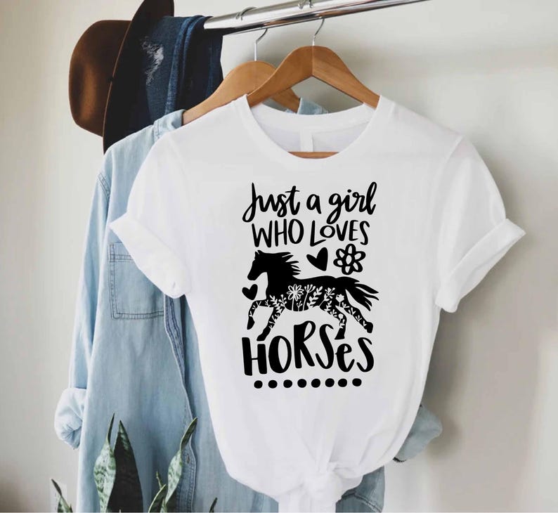 May include: White t-shirt with the text "Just a girl WHO LOVES HORSES" and a black horse graphic. The horse graphic has a floral design on its body. The shirt is hanging on a wooden hanger.